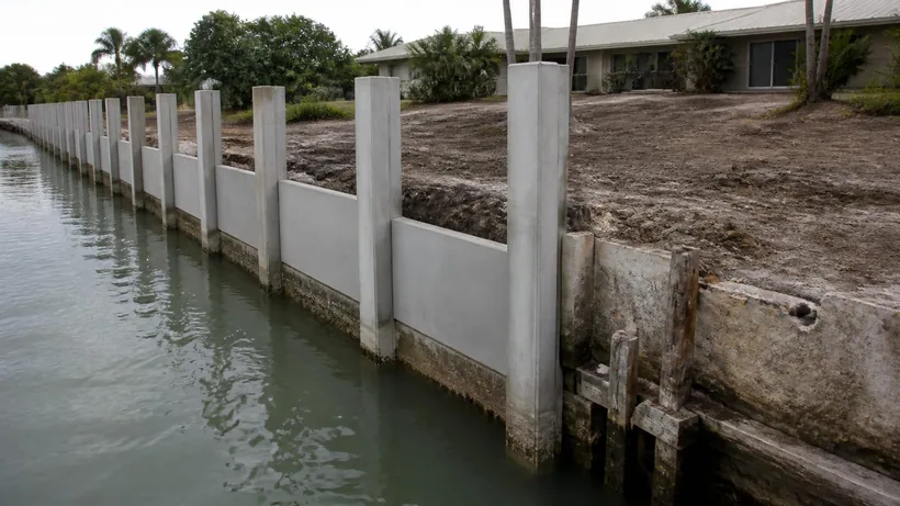 Newly driven concrete king piles standing along a canal edge beside the remains of the wall being replaced