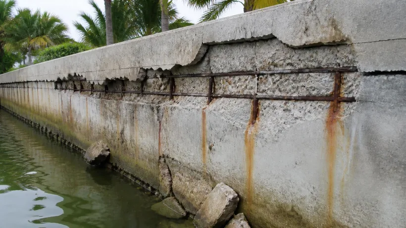 Concrete seawall face spalled along a band where corroded reinforcement has expanded, rust staining bleeding down the panels
