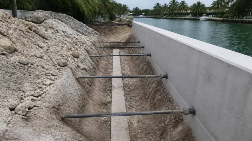 Open trench behind a seawall with horizontal steel tie rods running back to a buried concrete deadman anchor